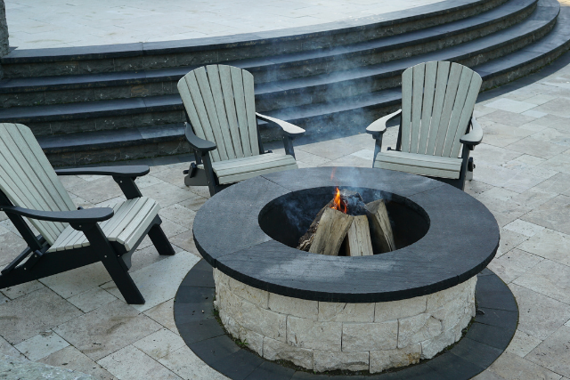 Round black sandstone fire pit with burning wood and Adirondack chairs on a patio