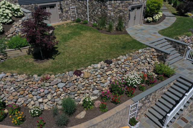 Multi-color river rock landscape bed with stone steps, retaining wall, and floral plantings at a residential property