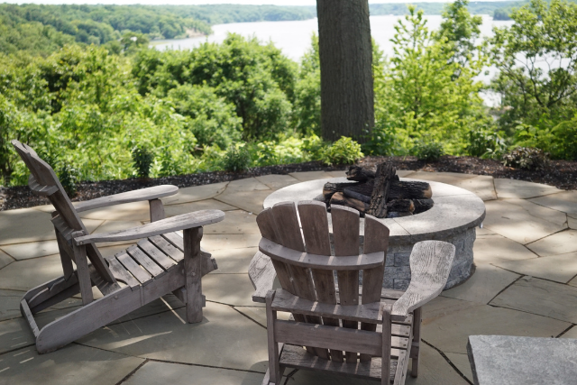 Bluestone flagstone patio with circular stone fire pit and Adirondack chairs overlooking a lake view