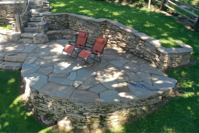 Round flagstone patio with stacked stone seating wall, steps, and lounge chairs in a backyard