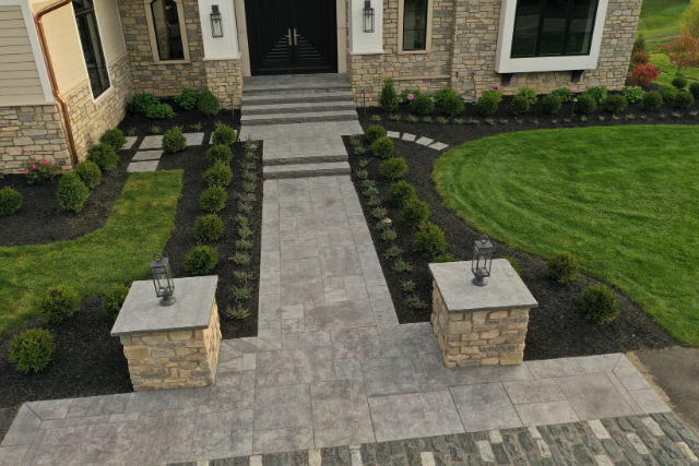 Grand stone walkway with pattern cut pavers, lantern-topped pillars, and manicured landscaping leading to a home entrance