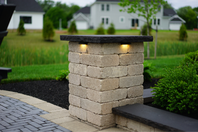 Stacked stone pillar with dark natural stone cap and inset lighting at a residential driveway entrance
