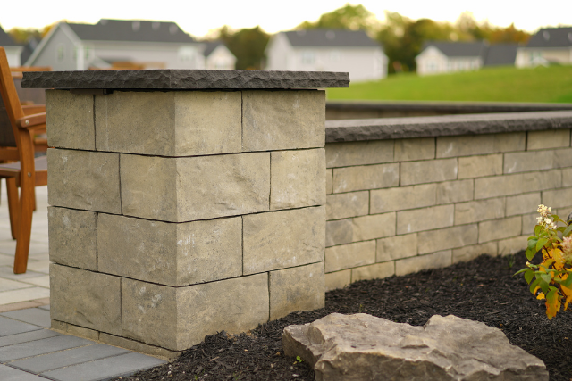 Tan stone block pillar with dark natural stone cap next to a matching wall in a residential landscape