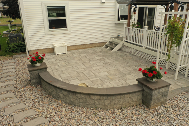 Curved paver patio with stone seating wall, pillar caps, and red flower planters at a residential home